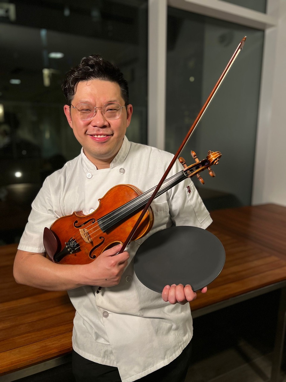 Abraham Wong smiling in a chef coat while holding a violin and a plate